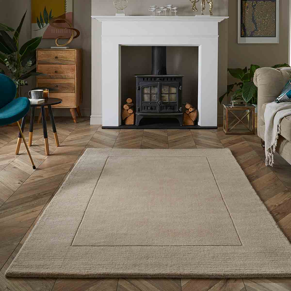 Beige rectangular area rug with a subtle border, placed on a herringbone wooden floor in a cozy living room featuring a black stove, green plants, and wooden furniture.