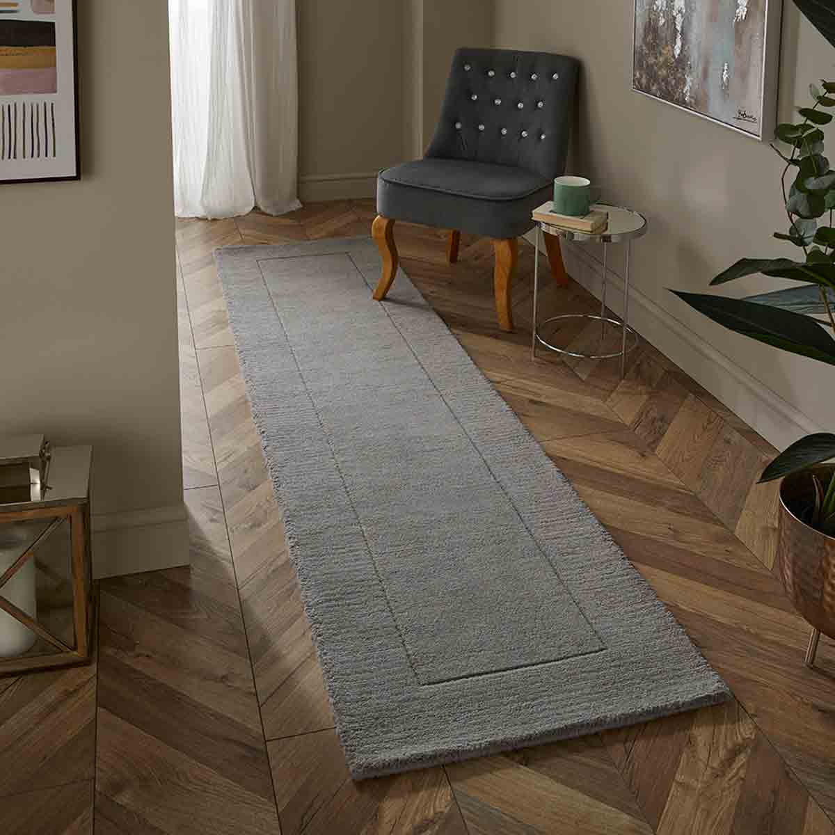 Mayfair Esme 60x230cm silver wool runner in grey, featuring a textured surface, bordered design, placed alongside a grey upholstered chair and a small side table.