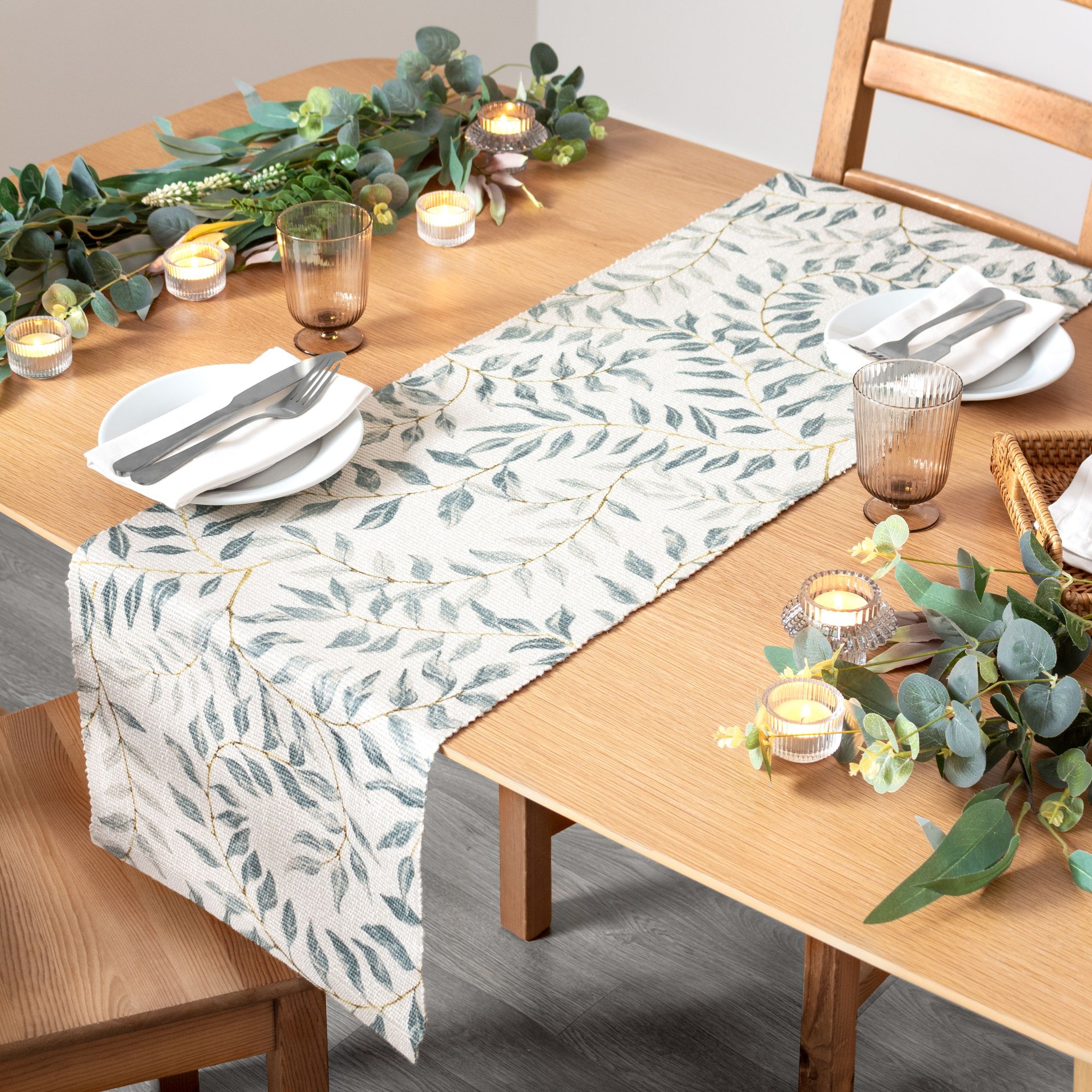 Large table runner featuring a light cream background adorned with green leaf patterns, made from durable fabric, perfect for indoor or outdoor use, styled on a wooden table.