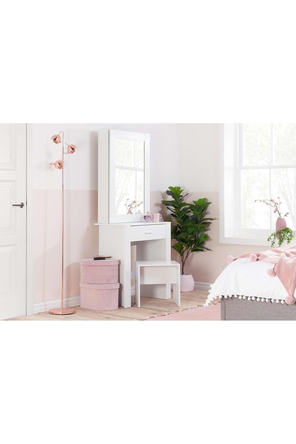 White wooden dressing table with a sliding mirror, featuring one drawer, accompanied by a pink stool, decorative plant, and a rose gold floor lamp in a softly colored room.