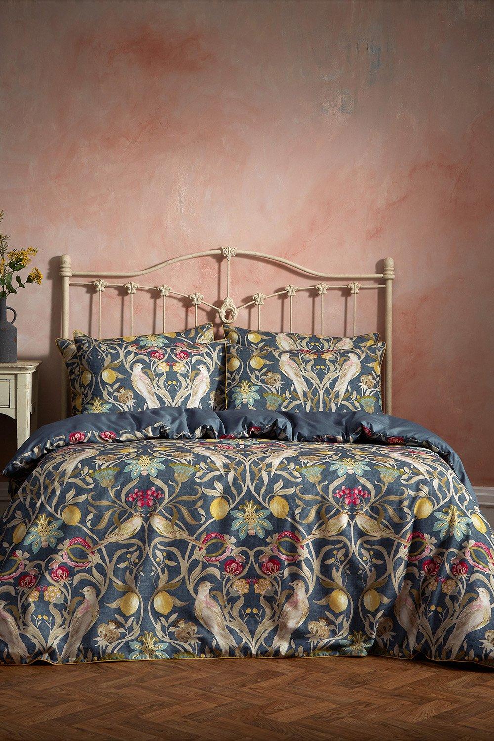 Luxurious navy cotton duvet cover set featuring intricate floral and bird patterns, complemented by matching pillowcases, set against a softly colored wall and vintage bed frame.