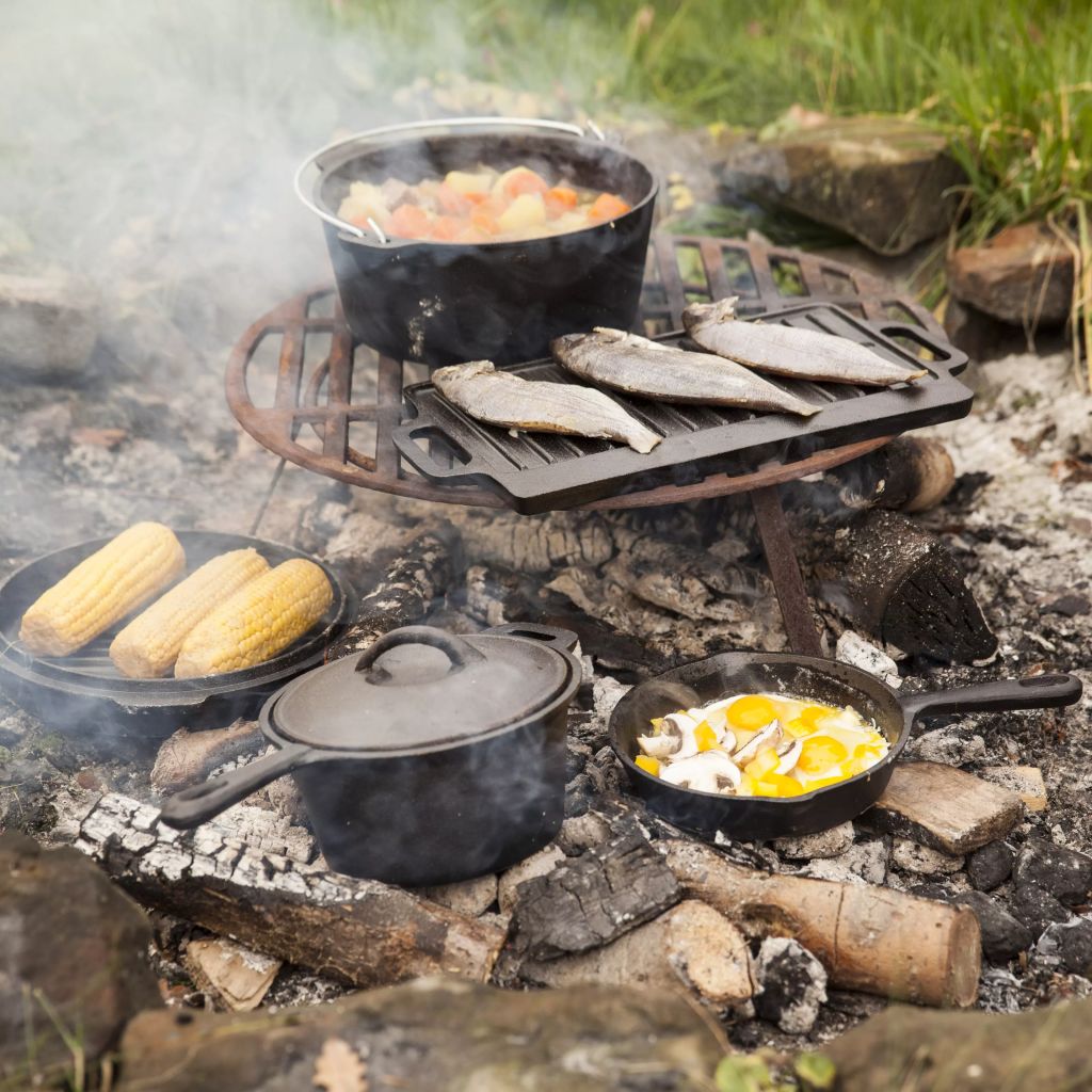 Seven-piece campfire cooking set featuring black cast iron pots and pans, with fish and vegetables cooking over an open flame on a metal grate amidst smoky embers.