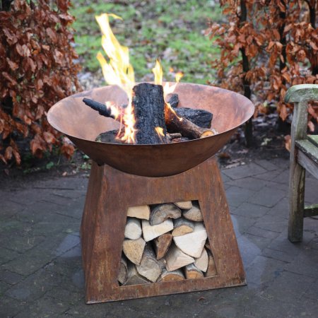 Round, rust-colored metal fire bowl with flames and logs, supported by a sturdy base that stores firewood, set on a stone patio surrounded by autumn foliage.