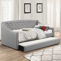 Gray fabric daybed with a curved back, tufted detailing, and a pull-out trundle, adorned with decorative pillows and a patterned throw blanket, set in a bright, airy room.