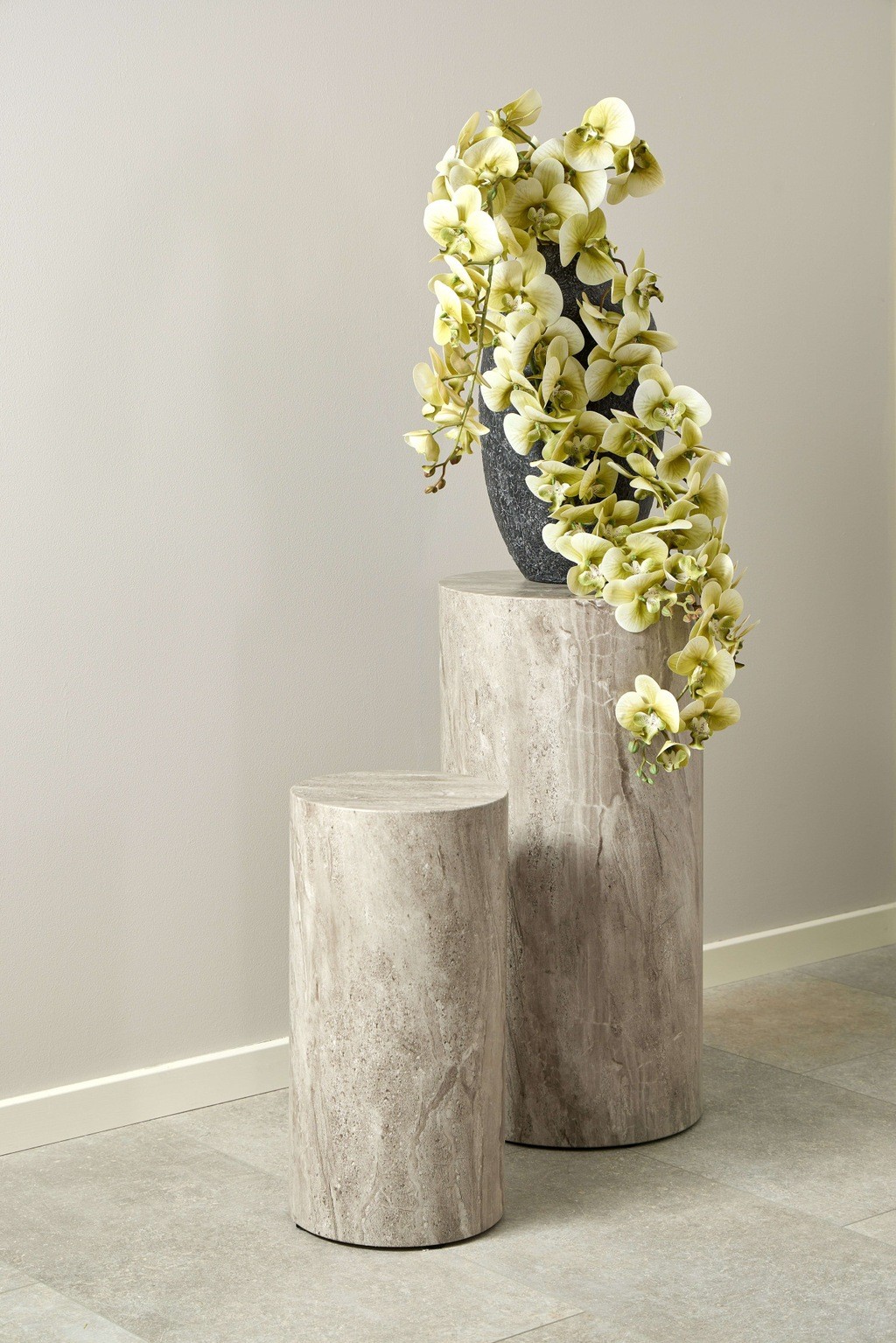 Set of two round pedestal tables in grey marble with a river rough paper finish, one taller and one shorter, adorned with a black vase and cascading yellow orchids.
