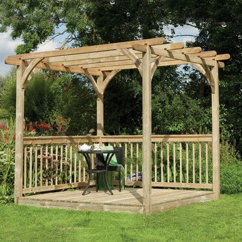 An 8' x 8' wooden pergola deck kit with a natural finish, featuring a slatted roof, arched supports, and a surrounding railing, set on a grassy area with greenery in the background.