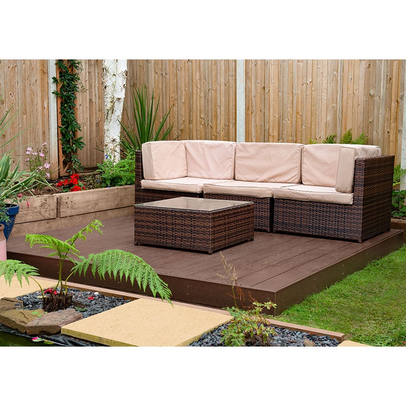 Brown composite decking kit measuring 8' x 8', featuring a smooth surface, surrounded by greenery, and complemented by a wicker sofa and coffee table with beige cushions.