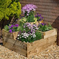 Tiered wooden raised bed in natural finish, measuring 3'x3', featuring vibrant purple and white flowers, set against a brick wall and gravel surface.