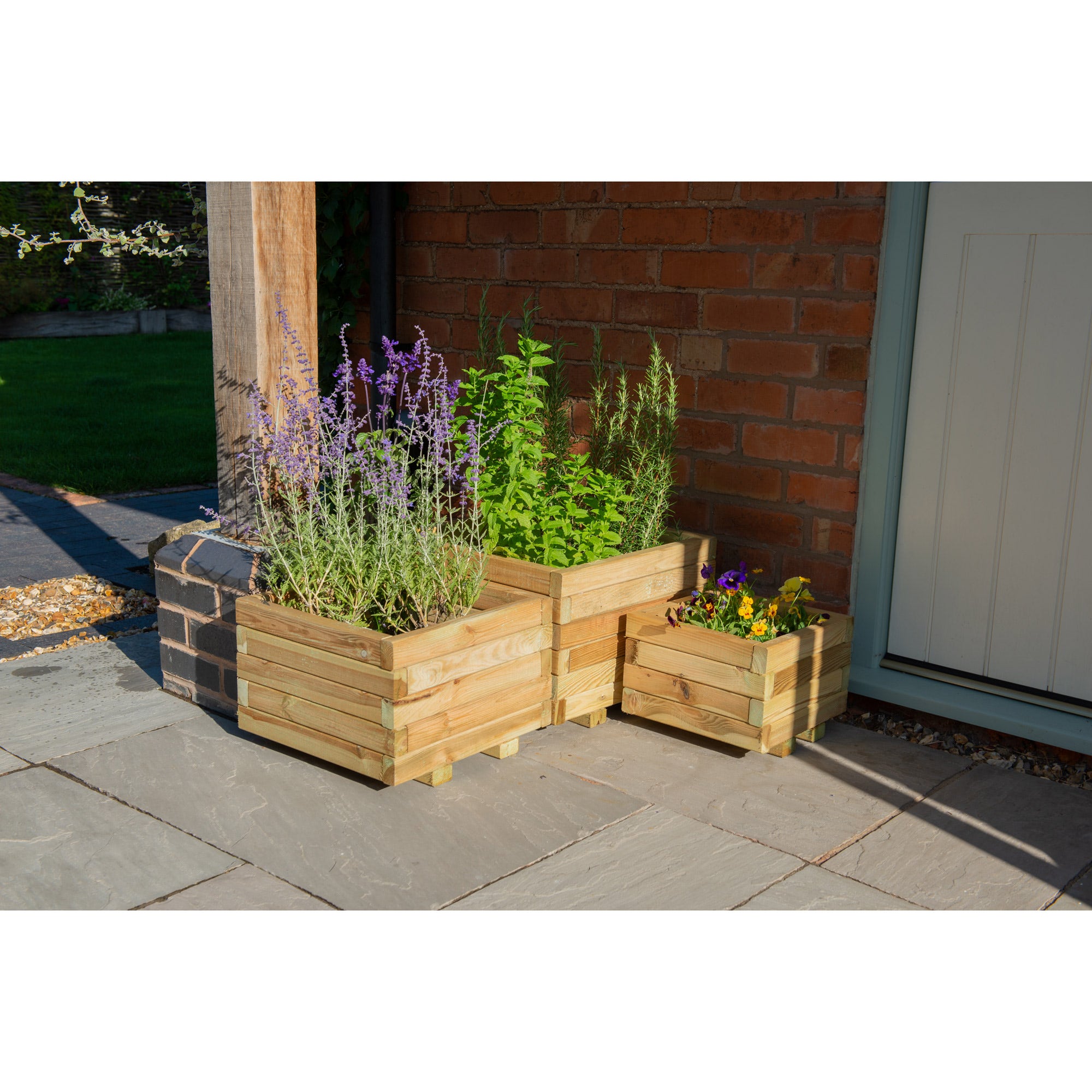 Square wooden garden planters in natural wood finish, featuring three varying sizes, filled with vibrant green plants and purple flowers, positioned on a stone patio.