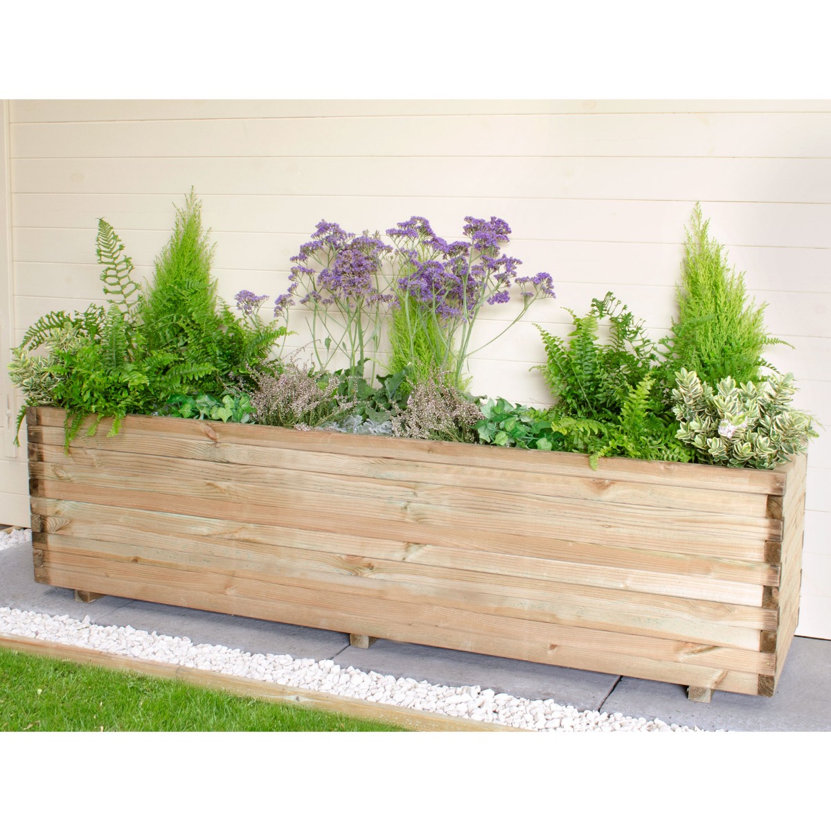 Wooden garden planter in natural wood finish, measuring 6'x2', filled with lush greenery and purple flowers, featuring a slatted design and sturdy feet on a stone pathway.