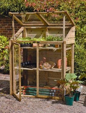 A small wooden lean-to greenhouse with a natural finish, featuring a slanted roof, glass panels, open front door, and shelves holding potted plants and gardening supplies.
