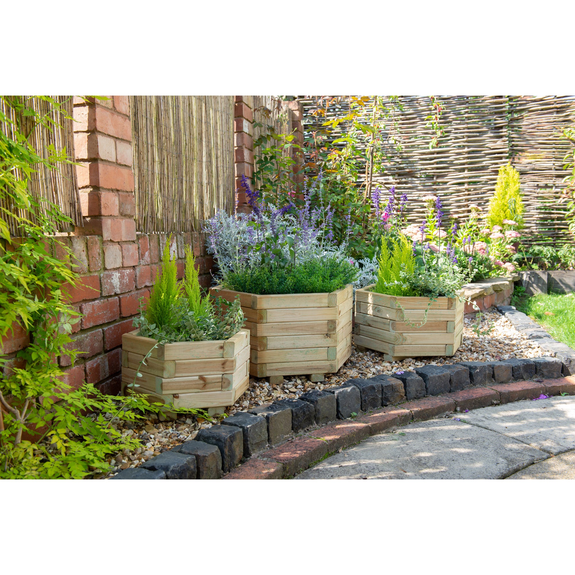 Three hexagonal wooden garden planters in natural wood finish, filled with vibrant green plants and purple flowers, positioned against a brick wall and stone pathway.