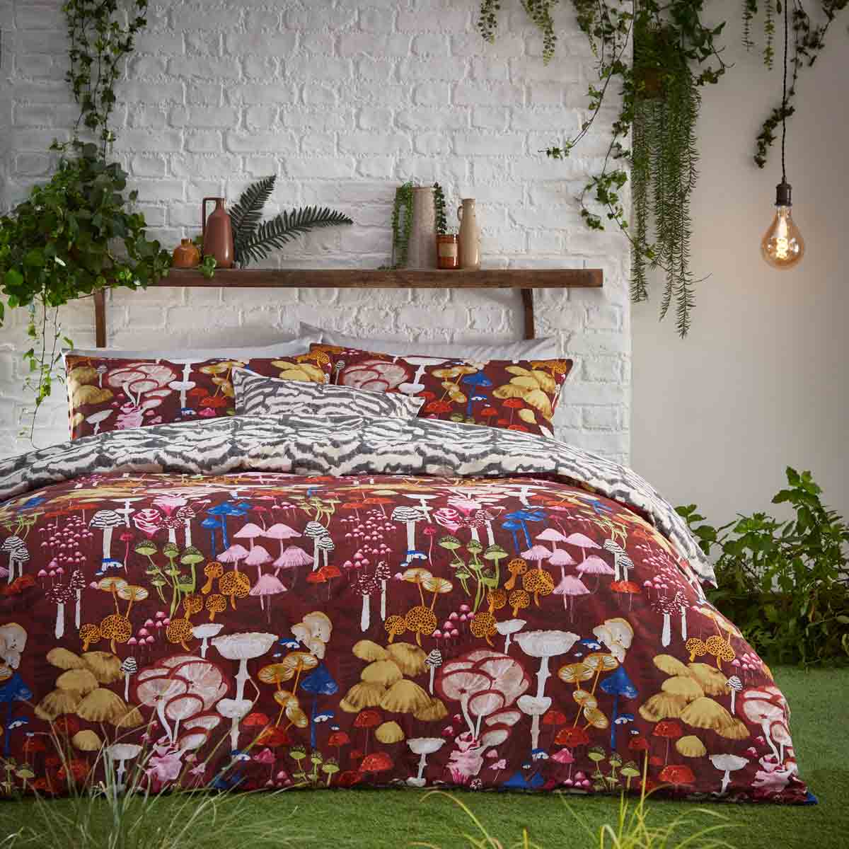 Red duvet cover set featuring abstract mushroom designs in vibrant colors, complemented by patterned pillowcases, set against a textured white brick wall and greenery.