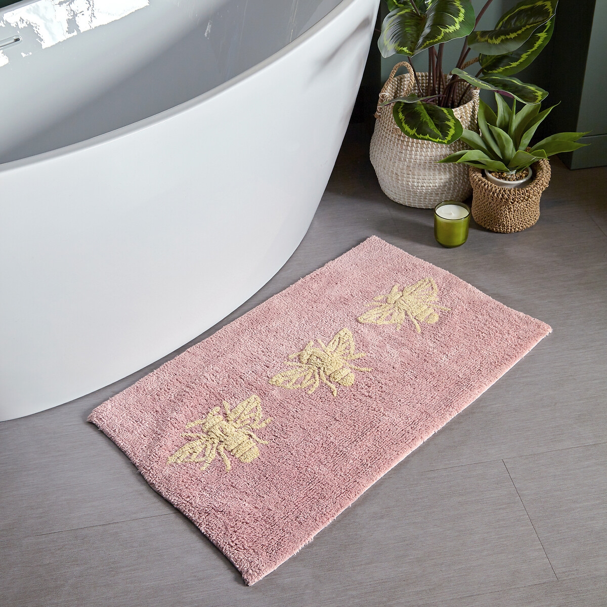 Blush cotton tufted bath mat featuring three decorative bees, non-slip backing, placed beside a modern bathtub with greenery in the background.