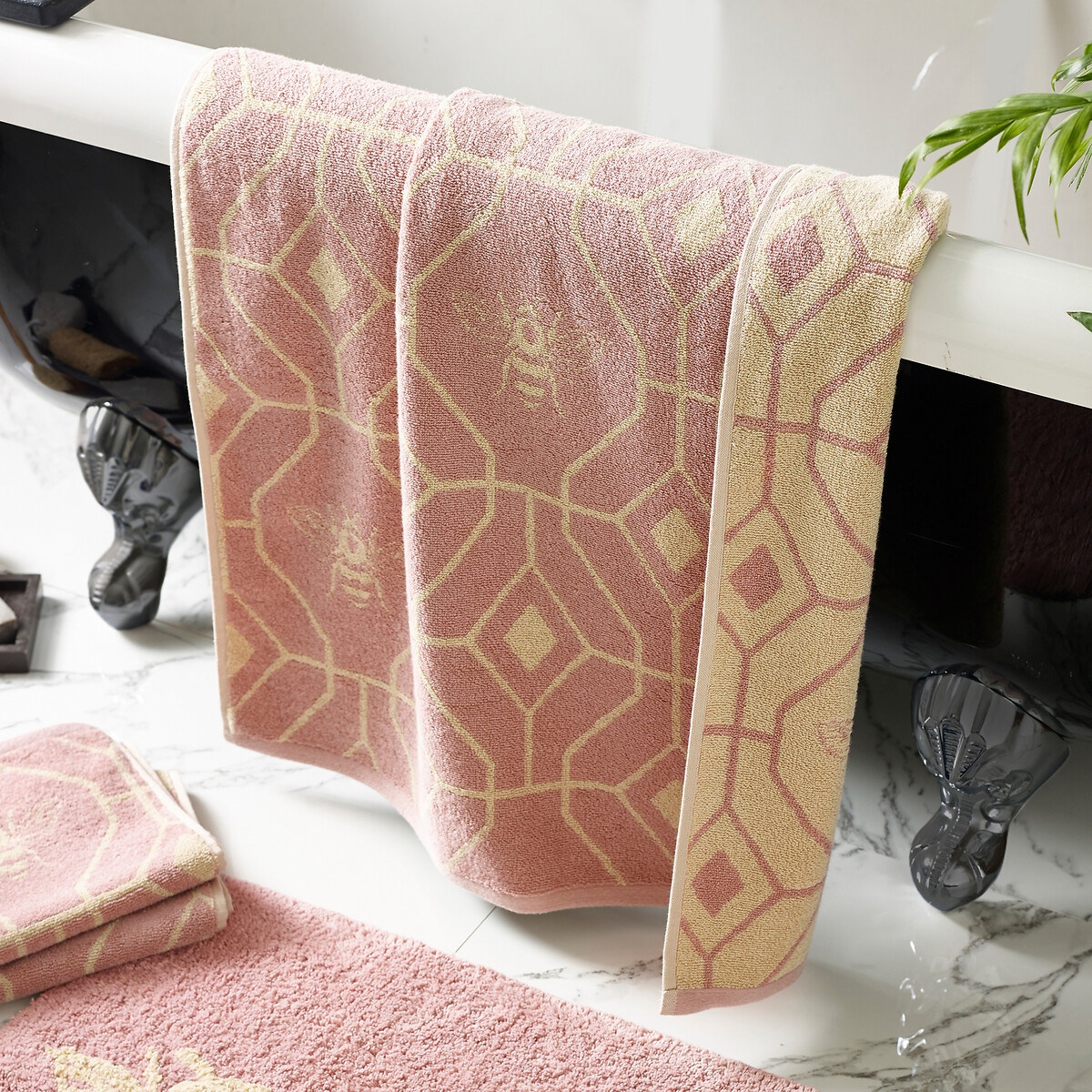 Geometric cotton jacquard hand towel in soft pink with a honeycomb pattern and bee motifs, featuring a beige border, elegantly draped over a bathtub.