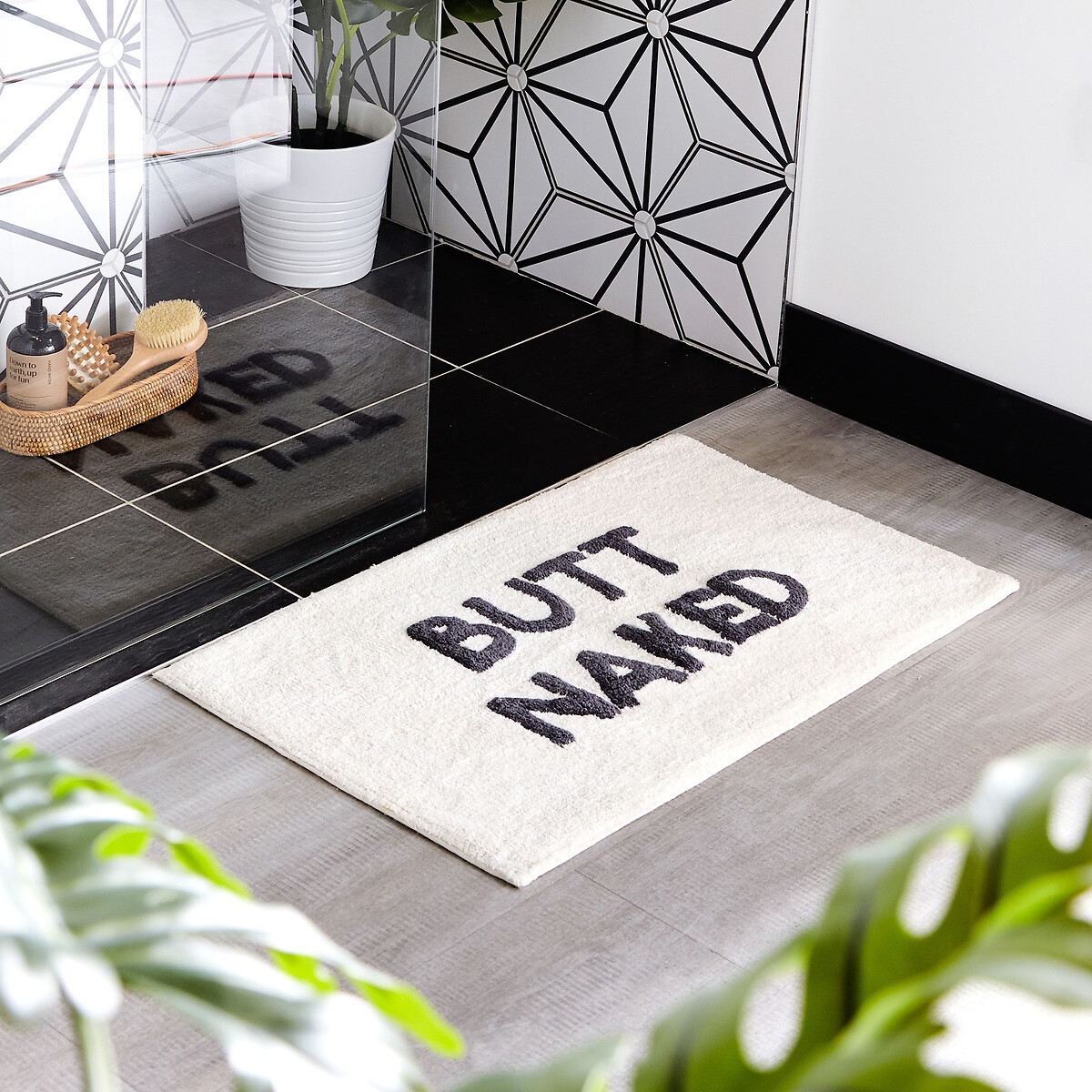 Ivory cotton tufted bath mat featuring the slogan "BUTT NAKED" in bold black letters, with a non-slip backing, placed on a tiled floor near a plant and decor.