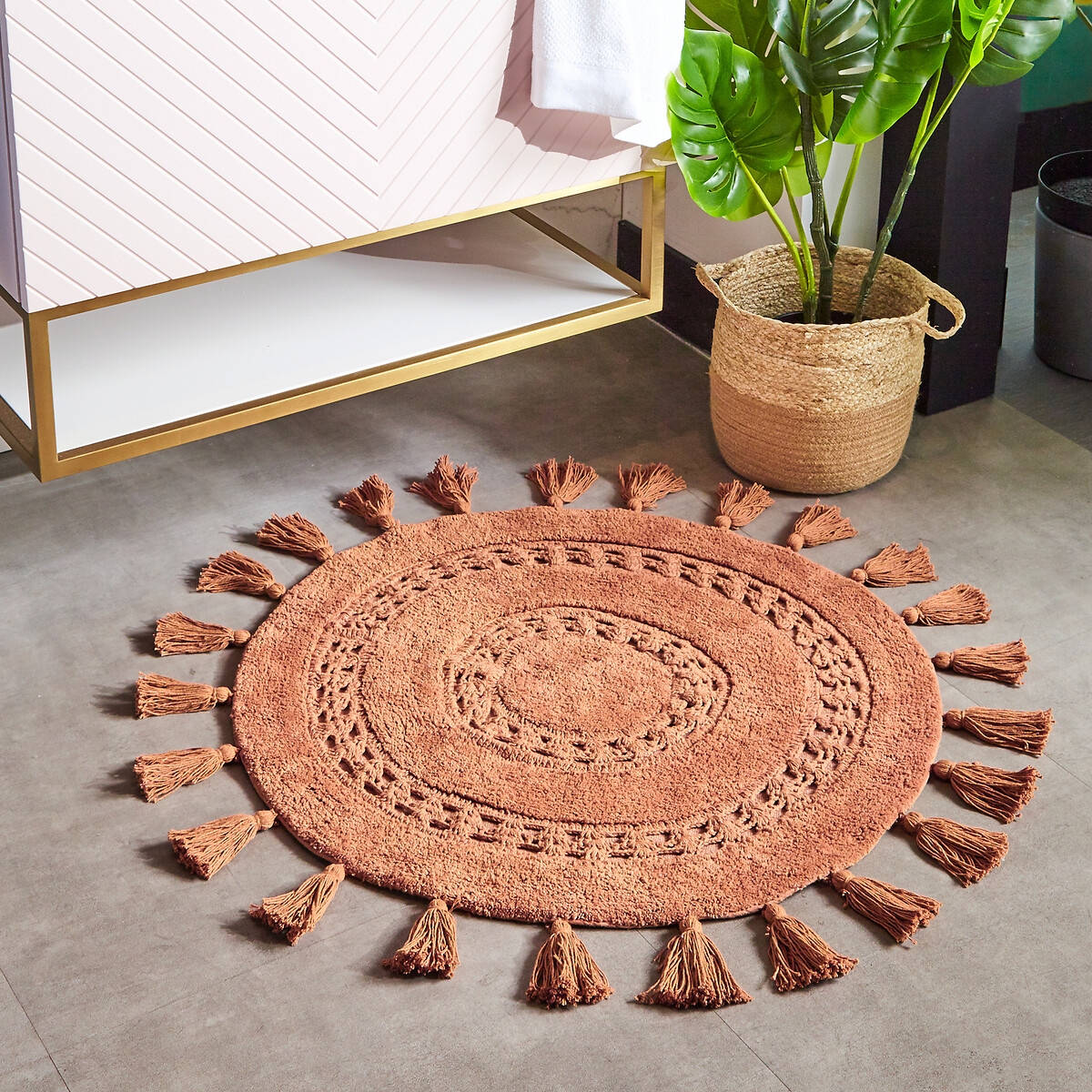 Round brown cotton bath mat featuring woven patterns and decorative tassels around the edge, placed on a tiled floor beside a potted plant and a modern vanity.