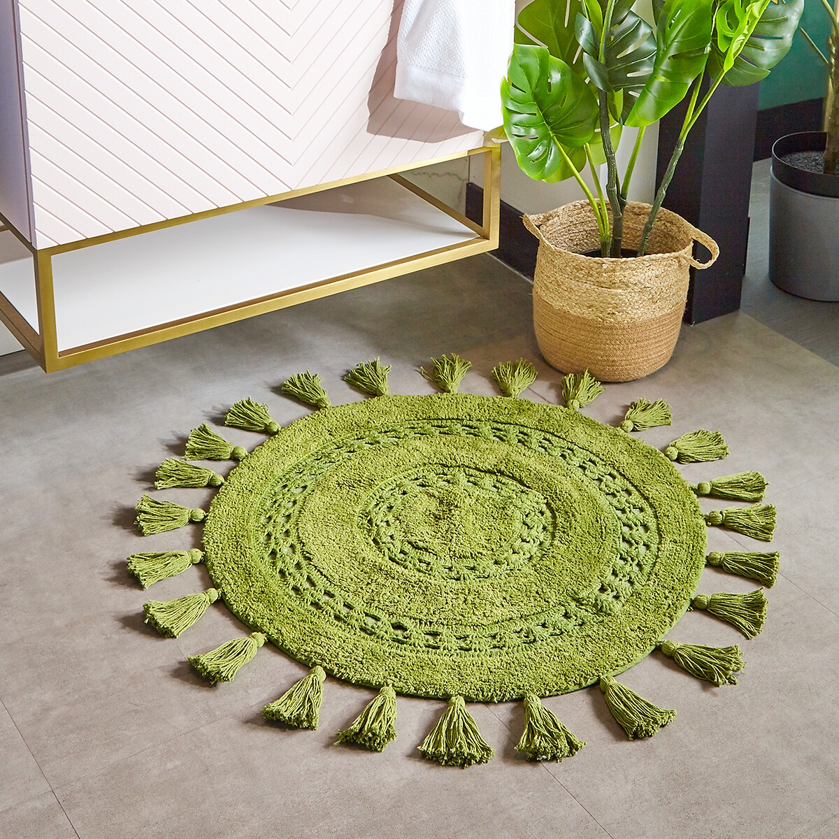 Round green cotton bath mat with woven texture and decorative tassels around the edge, placed on a tiled floor next to a plant and a modern cabinet.