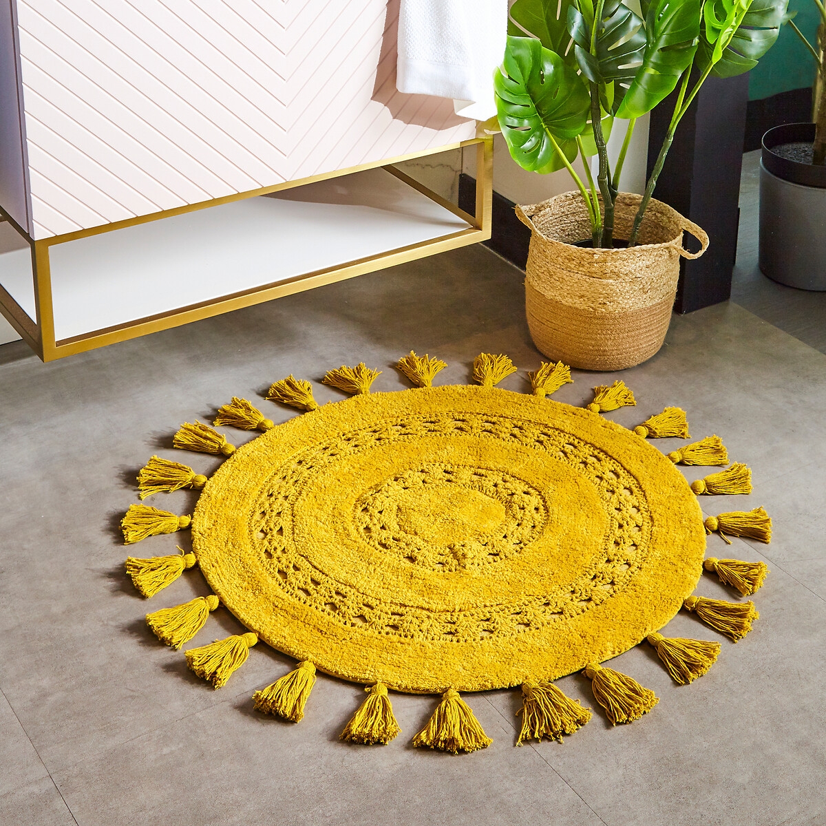Round yellow cotton bath mat with woven texture and decorative tassels around the edge, placed on a tiled floor near a plant and modern furniture.