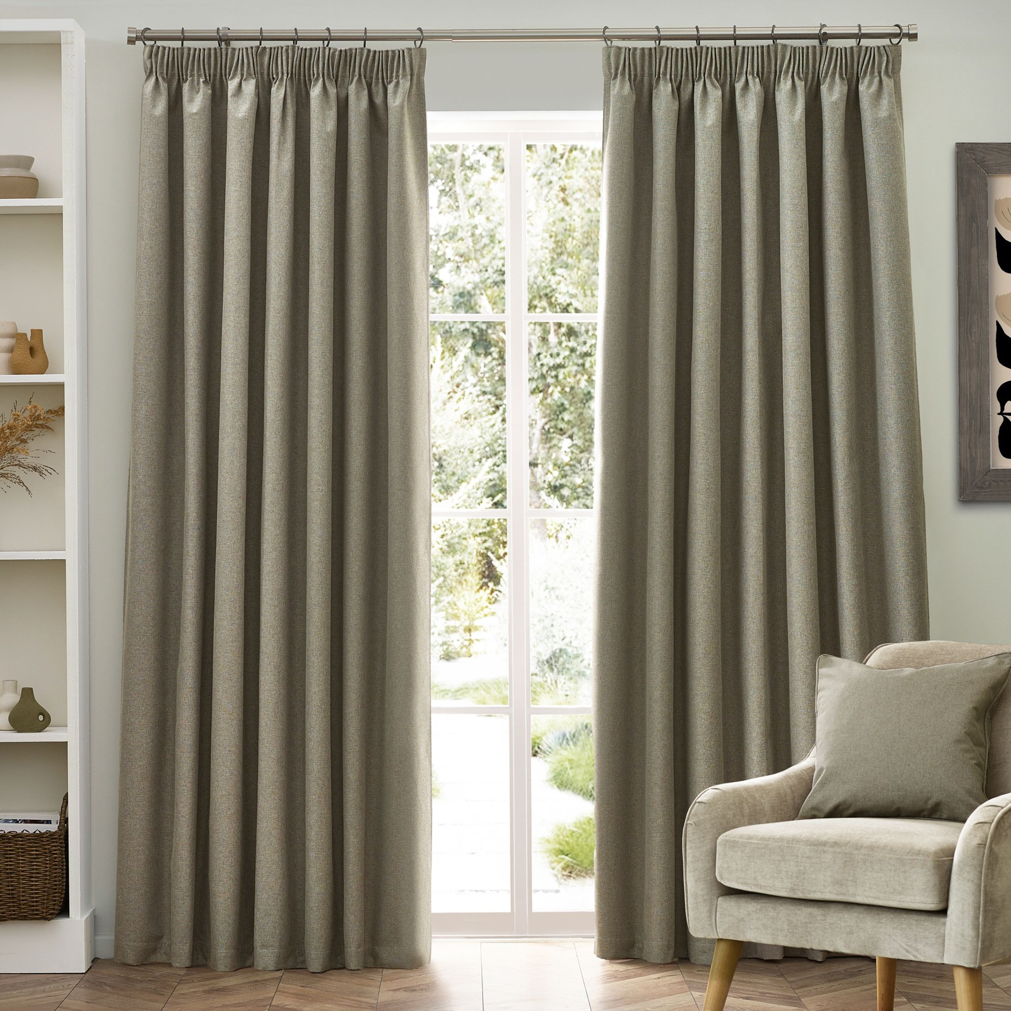 Natural-colored blackout pencil pleat curtains, measuring 117cm width by 137cm drop, hang elegantly by a window, complementing a light, airy room with a cozy armchair.