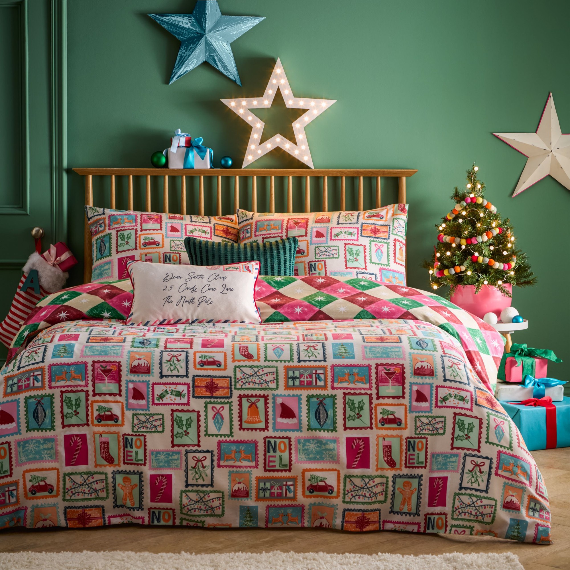 Super King duvet cover set featuring a colorful "Dear Santa" print with festive motifs, complemented by decorative pillows, against a green wall and a small Christmas tree.