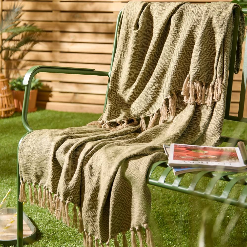 Olive green woven throw blanket with fringe edges draped over a green metal chair, set against a backdrop of wooden slats and greenery.