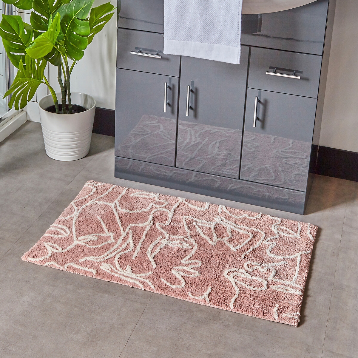 Abstract cotton tufted bath mat in blush pink with a non-slip backing, featuring a wavy white design, placed on a tiled bathroom floor near a plant and vanity.