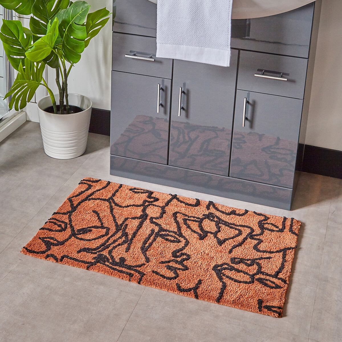 Brown abstract cotton tufted bath mat with non-slip backing, featuring a wavy black design, placed on a tiled bathroom floor near a potted plant and a vanity.