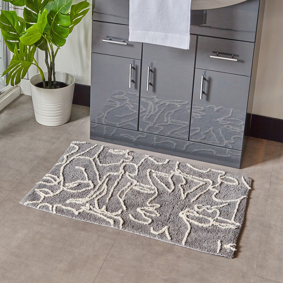 Abstract grey cotton tufted bath mat featuring a non-slip backing, with a textured design in lighter shades, placed on a tiled floor near a plant and bathroom vanity.