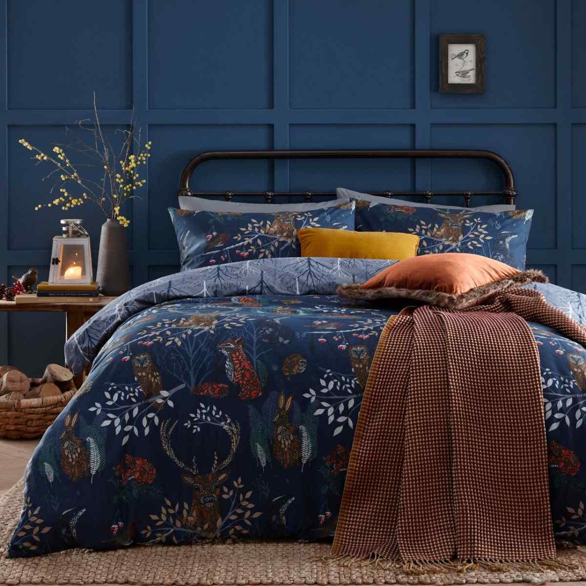 Navy reversible duvet cover set featuring woodland animals and foliage, complemented by textured pillows and a cozy throw, set against a blue paneled wall.