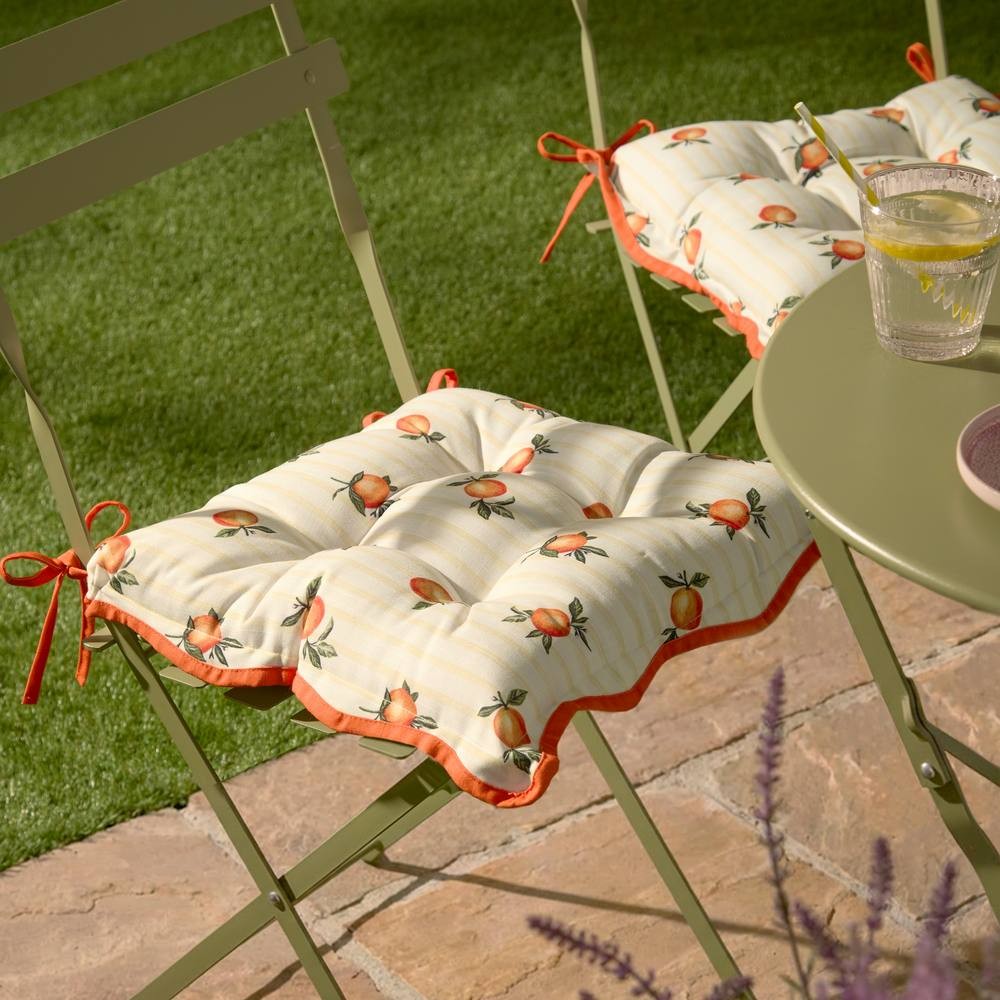 Yellow gingham seat pads featuring orange fruit patterns, with tufted design and ties for attachment, suitable for indoor or outdoor use, placed on green metal chairs.
