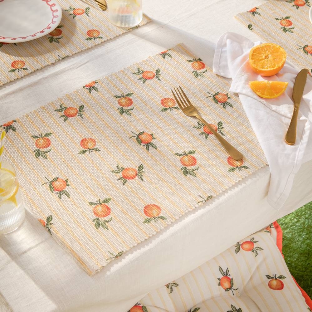 Set of 4 indoor/outdoor placemats featuring a natural fabric with orange fruit patterns on a striped yellow background, complemented by gold utensils and a fresh orange slice.