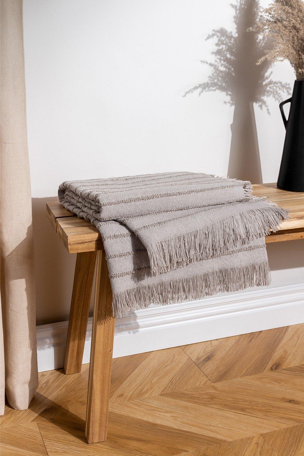 Light grey woven throw blanket with fringed edges, made from yarn, neatly folded on a wooden bench beside a black vase and dried flowers.