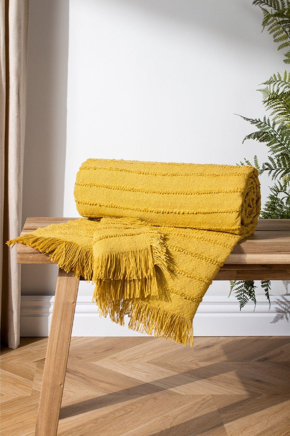 Yellow woven throw blanket with linear patterns and fringed edges, neatly rolled and partially draped over a wooden bench, with a backdrop of light-colored walls and greenery.