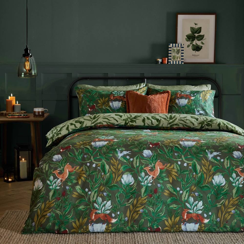 Reversible duvet cover set featuring a vibrant blue woodland print with animals and plants, complemented by green pillowcases and an orange accent cushion on a bed against a dark wall.