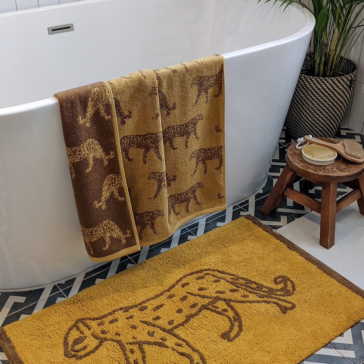 Leopard-patterned jacquard hand towel in gold and brown cotton, hanging on a white bathtub, with a matching bath mat on a patterned floor.