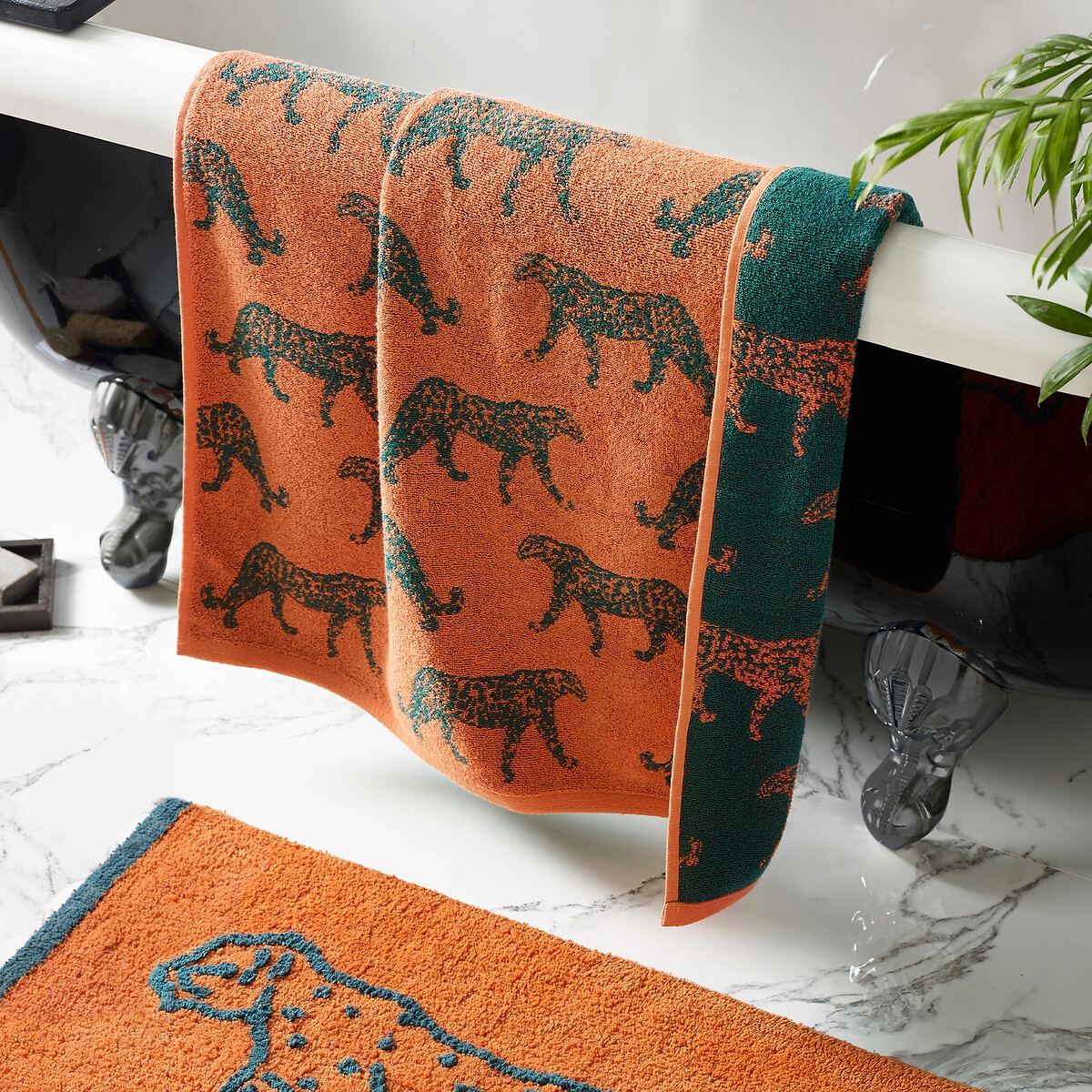 Orange cotton hand towel featuring a jacquard design of leopards in dark green, displayed hanging over a bathtub with a marble surface.