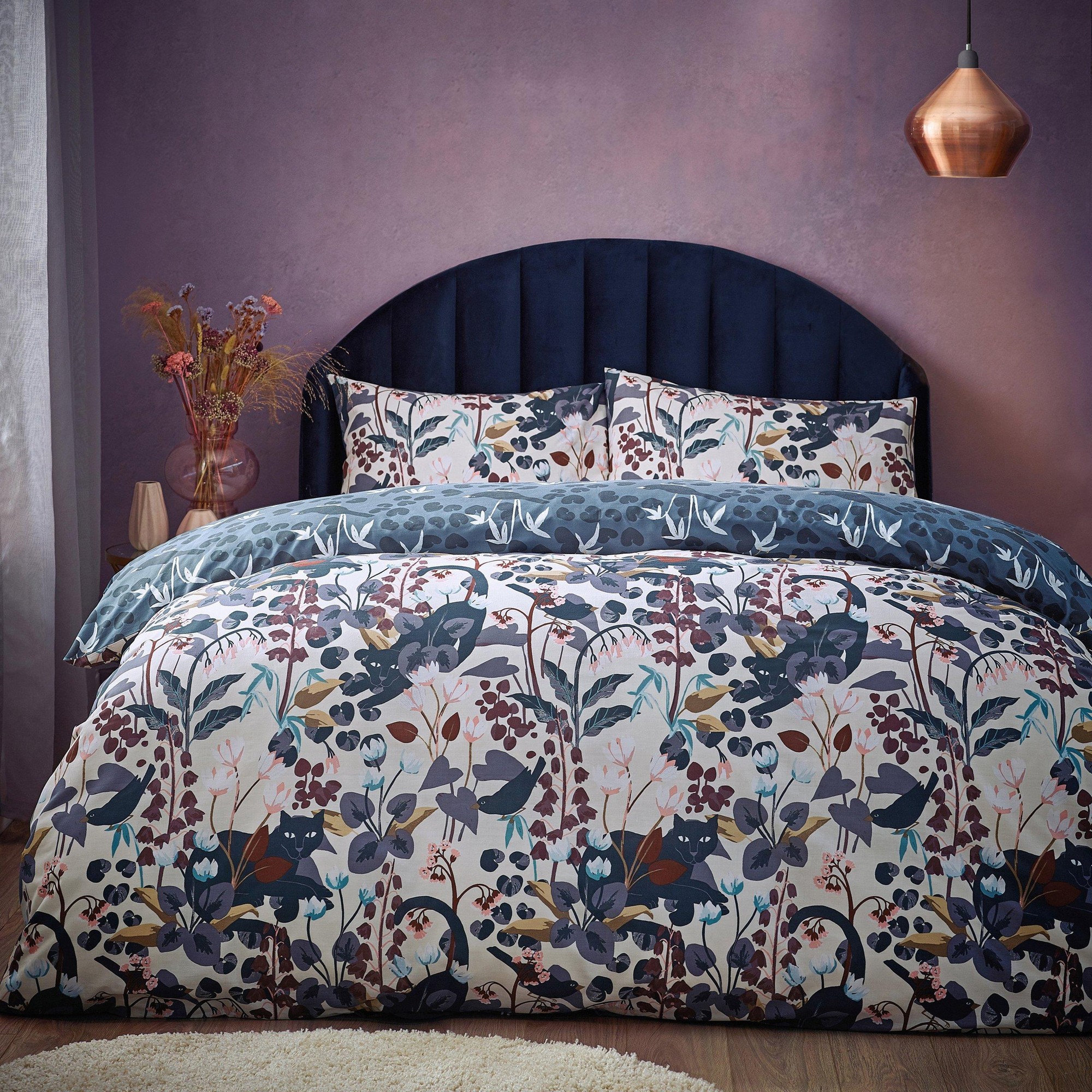 Floral-patterned bedding set featuring dark blue and multicolored botanical designs, complemented by a plush navy headboard and a copper pendant light.