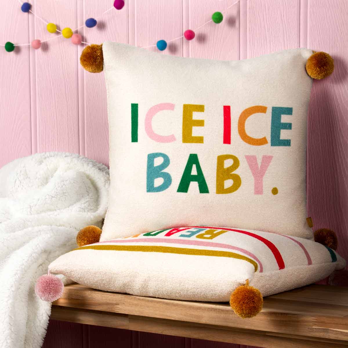Multicoloured cushion featuring the phrase "ICE ICE BABY" in bold letters, with fluffy pom-pom corners and a soft, textured fabric, set against a pink backdrop.