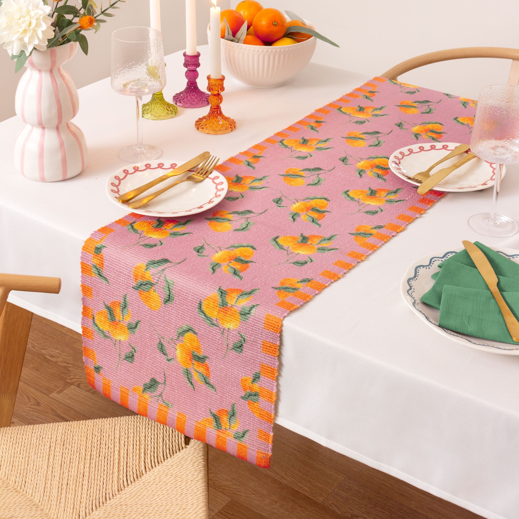 Pink indoor/outdoor table runner featuring vibrant orange fruit patterns, bordered with orange stripes, placed on a white table with decorative plates and glasses.