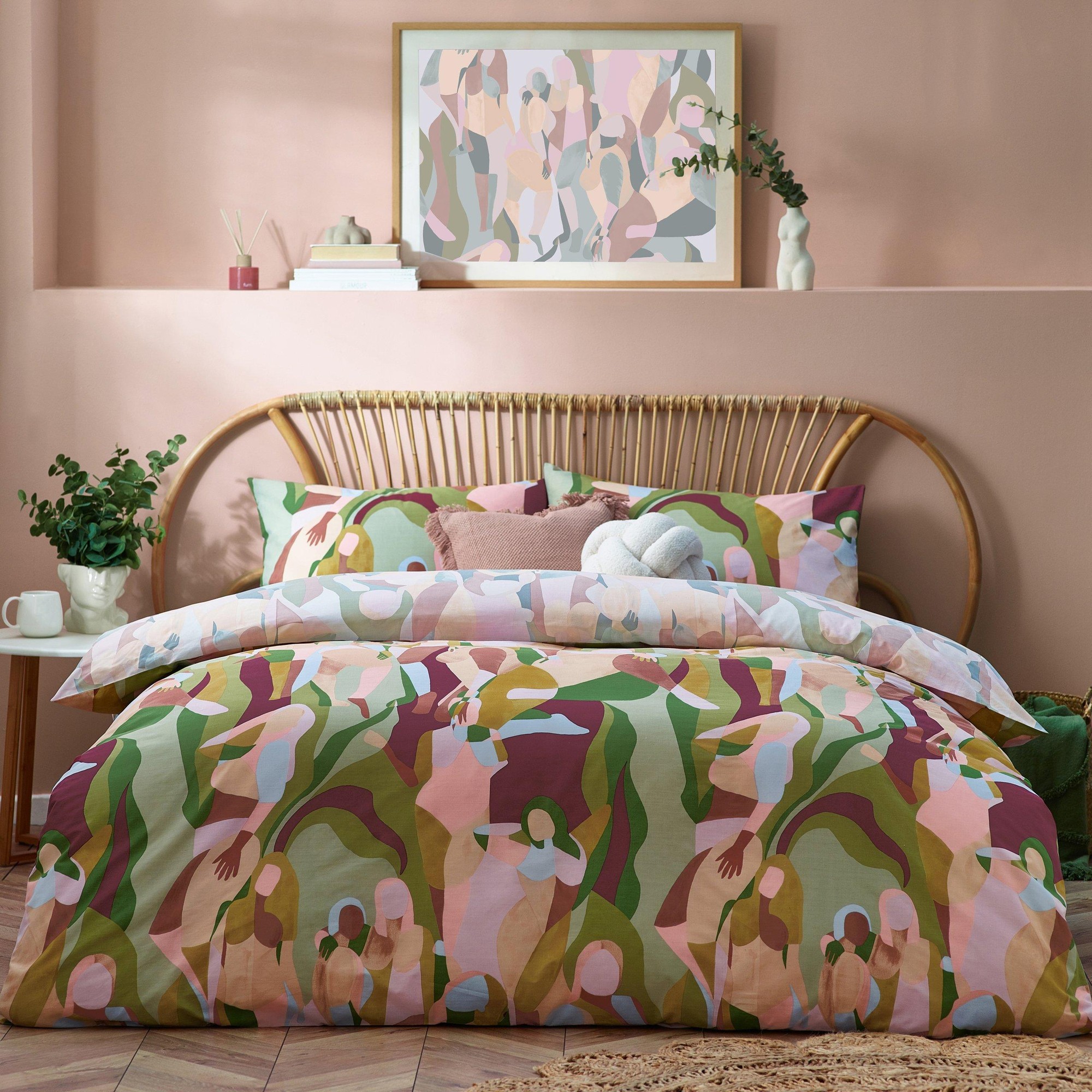 Multicolour abstract duvet cover set in polycotton featuring stylized figures, complemented by coordinating pillows, set against a soft pink wall and rattan bed frame.