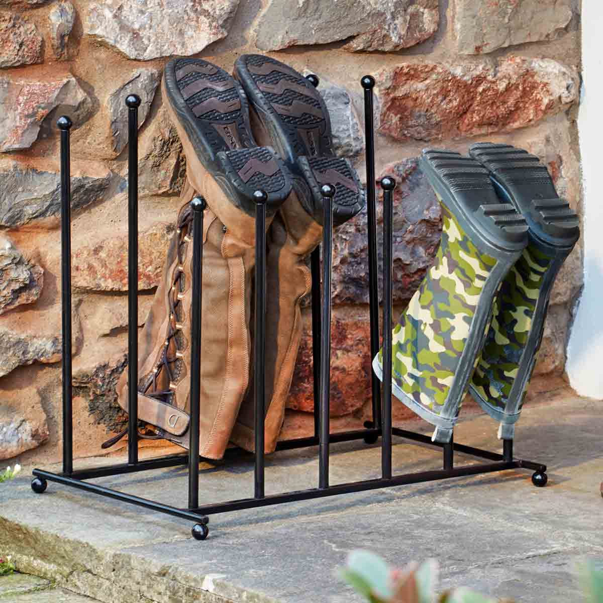 Black metal two-tier boot stand holding six pairs of boots, featuring sturdy rods and a sleek design, placed against a stone wall with brown and camo-patterned footwear.