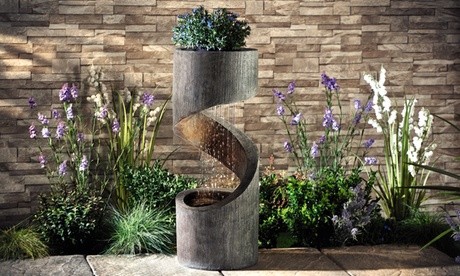Spiral-shaped water feature in gray stone finish, featuring a top planter, cascading water, and integrated LED lights, surrounded by colorful flowers and lush greenery.