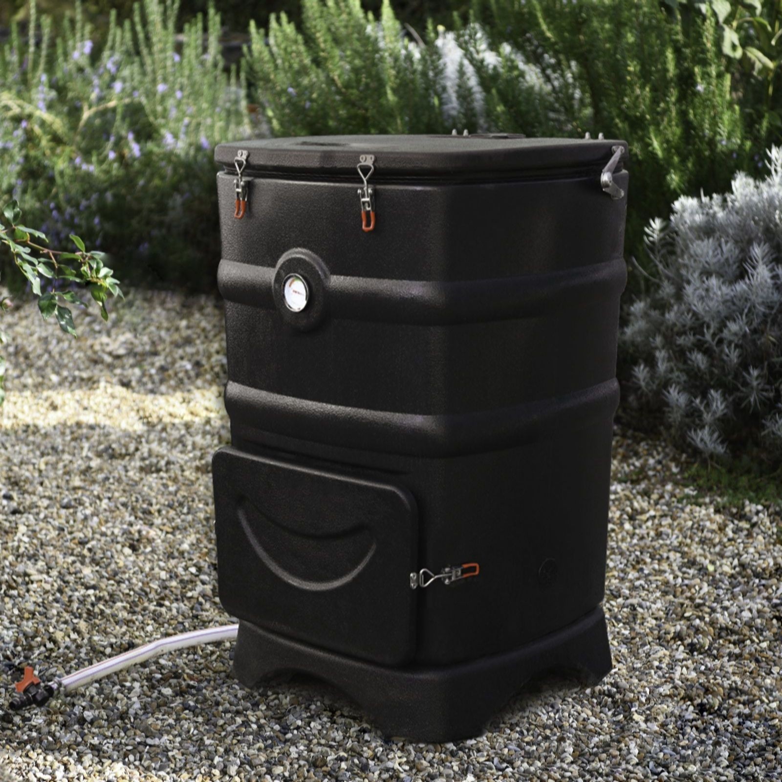 Black plastic compost bin with a lid, featuring metal clamps, a front access door, and a thermometer, set on gravel among green plants.