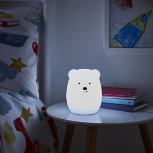 A soft, white bear-shaped night light with a friendly face, sitting on a wooden table beside colorful books and a patterned pillow featuring stars and shapes.