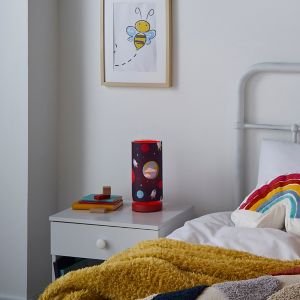 Red cylindrical table lamp with a space-themed design, featuring planets and stars, placed on a bedside table next to colorful books and a cozy blanket.