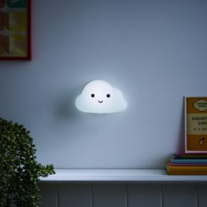 A soft, white cloud-shaped night light with a smiling face, featuring color-changing capabilities, mounted on a wall above a shelf with colorful books and a plant.