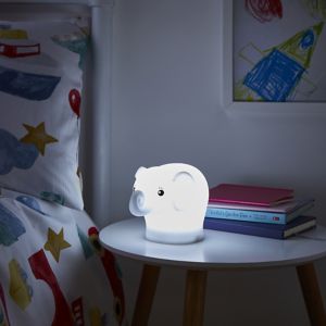 A cute, white elephant night light with a soft glow, made of plastic, sitting on a wooden table beside colorful books and a patterned pillow.