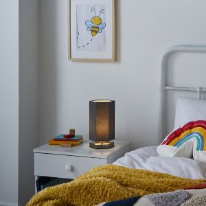 Silver hexagon table lamp with a textured fabric shade, illuminating a warm glow, placed on a bedside table next to a colorful throw and artwork featuring a bee.