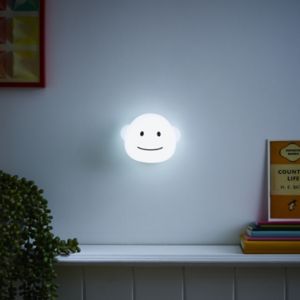 A round, white wall light shaped like a smiling monkey's face, emitting a soft glow, mounted on a light-colored wall with colorful books and framed art nearby.